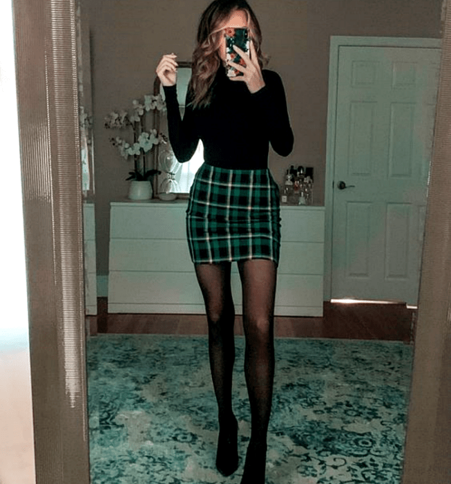 Chica de cabello claro usando un top negro de manga larga, minifalda verde de cuadros escocesa, medias negras semitransparentes y botines negros de tacón
