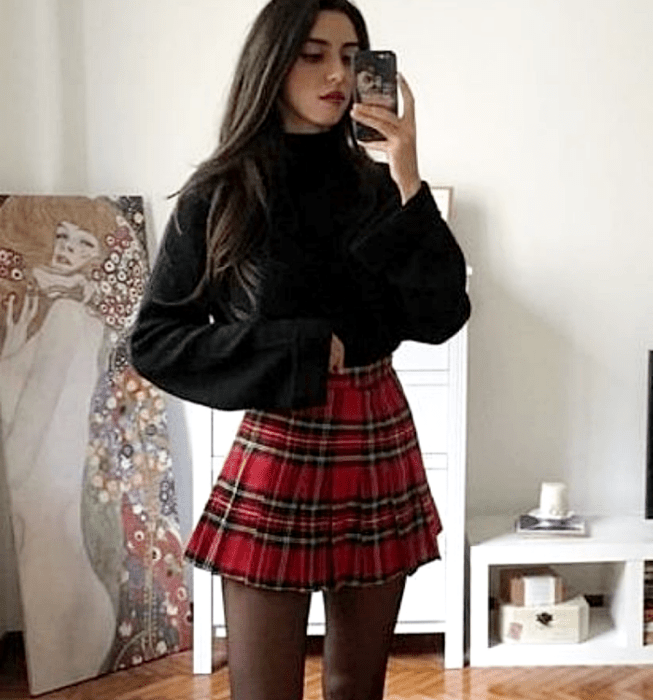 Chica de cabello castaño largo usando un suéter negro de manga larga y cuello alto, minifalda roja de cuadros con tablones al estilo escocés, medias negras semitransparentes