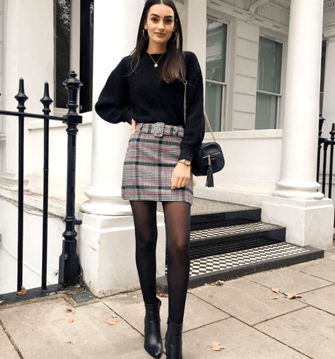 Chica de cabello castaño largo usando un suéter negro de manga larga, minifalda gris de cuadros escocesa, medias negras semitransparentes, botines de cuero de tacón y bolso de mano negro de cuero