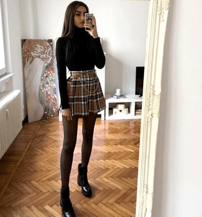 Chica de cabello castaño usando un top negro de manga larga y cuello alto, minifalda de tablones amarilla con cuadros estilo escocés, medias negras semitransparentes, botines negros de tacón
