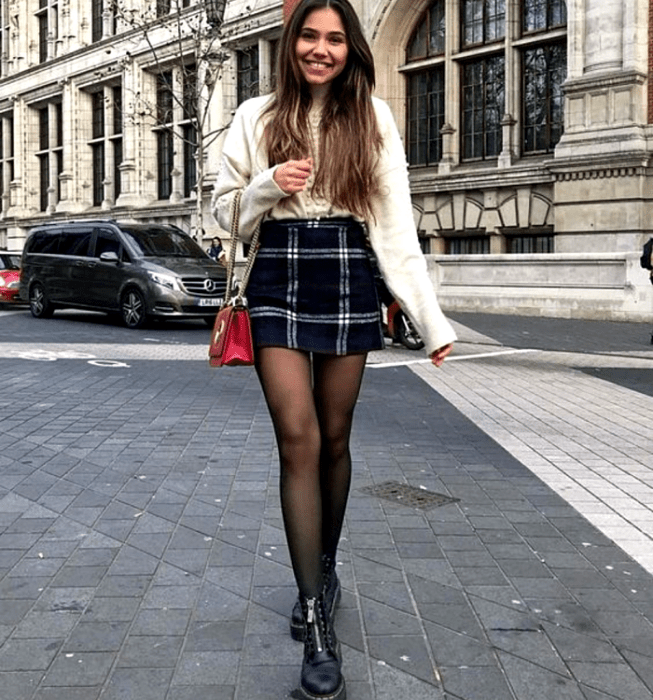 Chica de cabello castaño claro usando un suéter blanco de manga larga y cuello alto, minifalda azul marino con cuadros estilo escocés, medias negras semitransparentes, botines negros de suela gruesa y bolso mini de cuero rojo