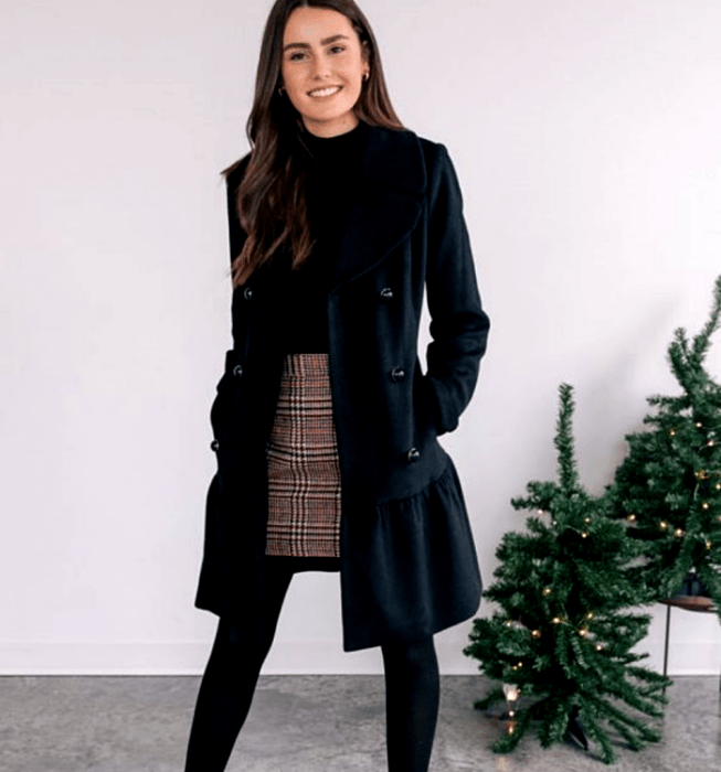 Chica de cabello castaño largo usando un top negro, abrigo largo azul marino, minifalda gris de cuadros estilo escocés, medias negras gruesas