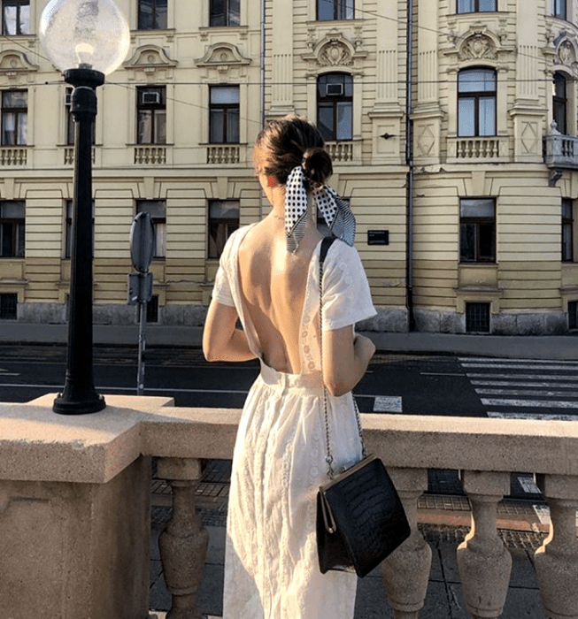 chica de cabello oscuro usando un vestido blanco de manga corta con escote en la espalda, bolso de mano negro con correa de cadena