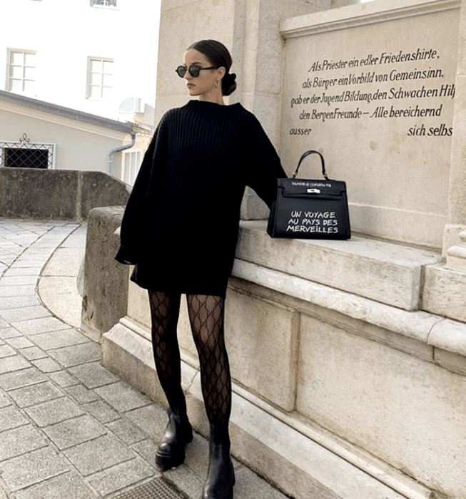 chica de cabello negro con lentes de sol, suéter oversized negro, minifalda negra, medias negras de red y botas de plataforma negras, bolso de mano negro con letras blancas