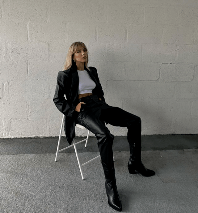 chica rubia usando un crop top blanco, abrigo largo de cuero, pantalones de cuero negros, botines de cuero con tacón