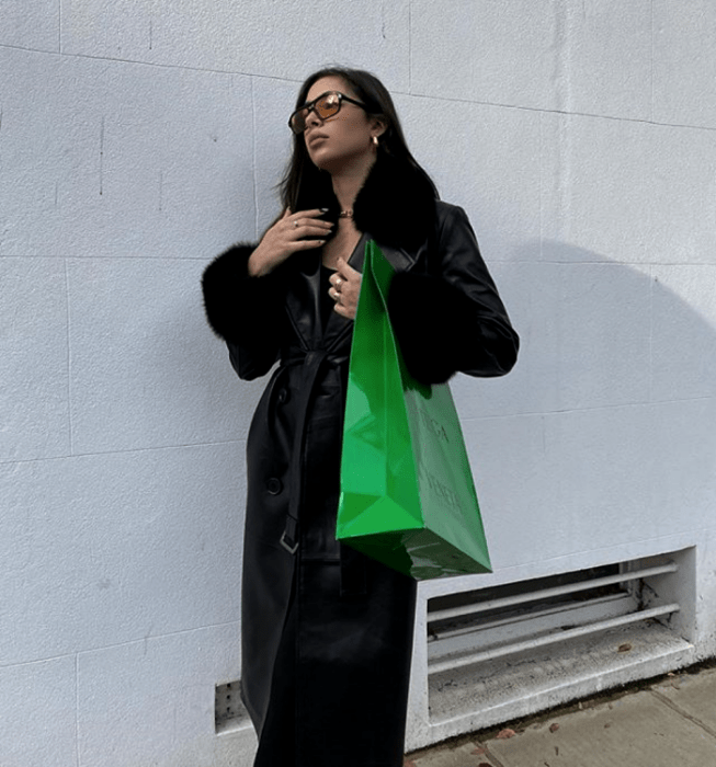 chica de cabello oscuro usando lentes de sol, abrigo largo de cuero negro con detalles de terciopelo en las solapas, bolso verde brillante