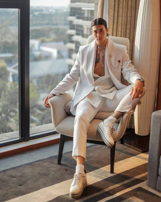 Paing Takhon sentado en un sillón con un traje blanco y zapatillas blancas