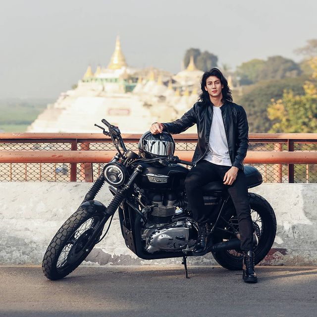 Paing Takhon posando en una motocicleta negra vistiendo pantalón y chaqueta negra con camiseta blanca 