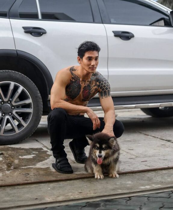 Paing Takhon en cuclillas con pantalón negro, pecho descubierto acariciando a un perrito malamute