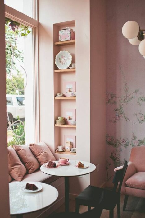 Cafetería rosa con un gran ventanal que da a la calle con asiento decorado con tres cojines rosa y una mesa redonda