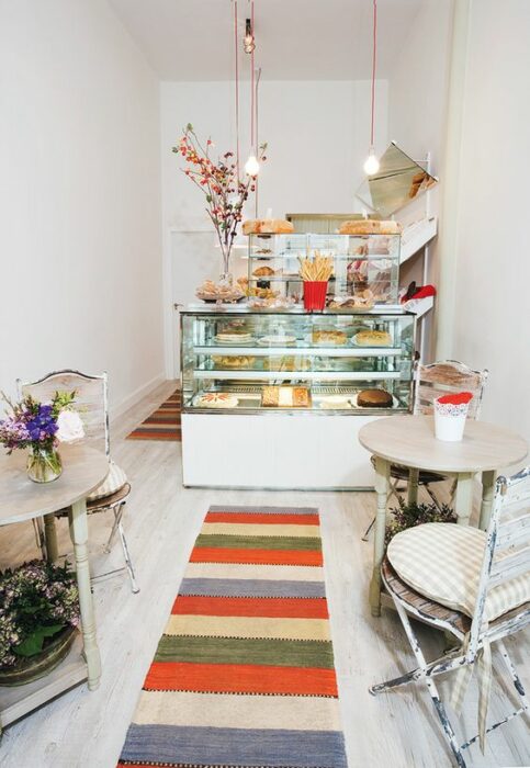 Cafetería blanca con mesas redondas beige y una alfombra colorida que conduce a una vitrina