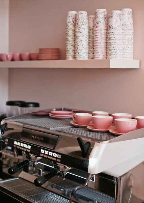 Cafetería con paredes rosas, platos y tazas rosas y una máquina de café plateada