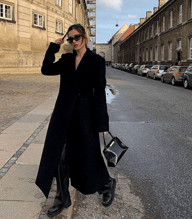 chica de cabello castaño usando lentes de sol, camiseta blanca, abrigo largo blanco, pantalones blancos de mezclilla, botines negros con punta beige y bolso negro de mano