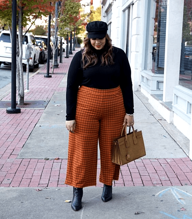 chica curvy de cabello castaño claro usando un gorro negro, suéter negro de cuello alto, pantalones naranjas con líneas negras, botines de piel negras de tacón y bolso café de mano