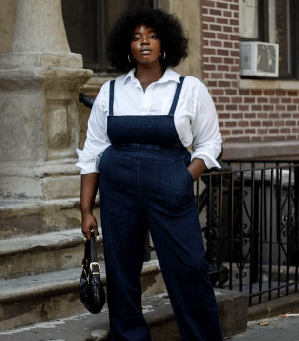 chica curvy morena de cabello chino usando una camisa de vestir blanca, overol de mezclilla, bolso negro de mano