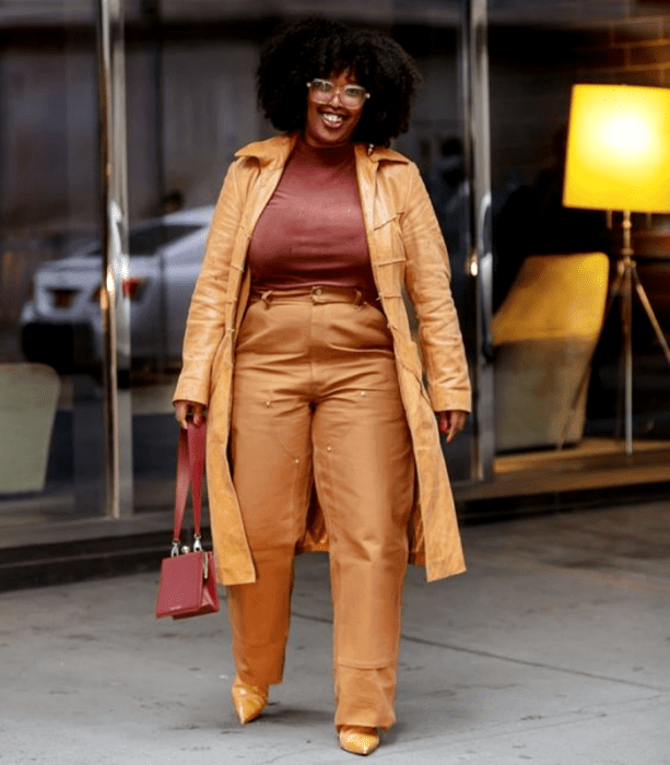 chica morena curvy de cabello chino usando un top café, gabardina beige de piel, pantalones de vestir beige de cuero y bolso café de mano