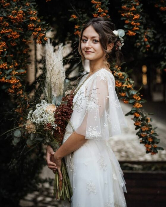 Mujer castaña con recogido de novia despeinado con vestido estilo bohemio con mangas transparentes y un ramo de flores en color guinda, blanco y naranja