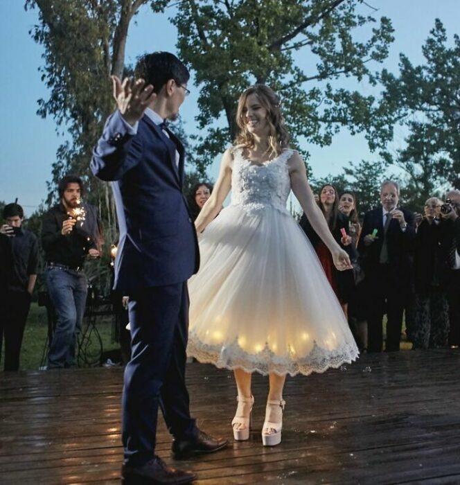 Pareja de novios bailando en la pista con los invitados mirándolos al rededor; él con traje azul marino y ella con vestido de encaje hasta el tobillo con luces en la falda