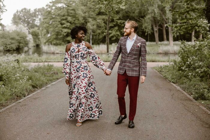Pareja de esposos caminando de la mano por una carretera en el bosque, ella con un vestido de novia largo, blanco con flores azules y ramas rojas con mangas largas y hombros descubiertos y él con un saco café con cuadros rojos, camisa blanca y pantalón guinfa