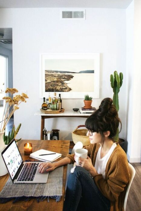 Chica trabajando en la computadora, su estudio tiene varias plantas, como un cactus o una platita pequeña cerca de la computadora