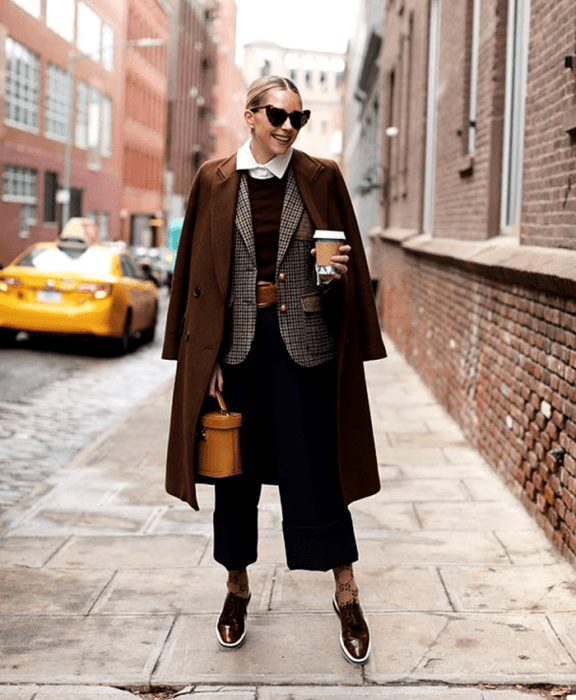 chica rubia usando lentes de sol en forma de corazón, camisa de vestir de cuello, suéter azul marino, cinturón café, saco café de cuadros, abrigo café largo, pantalones de vestir negros, calcetas cafés, zapatos cafés de vestir, bolso bucket naranja