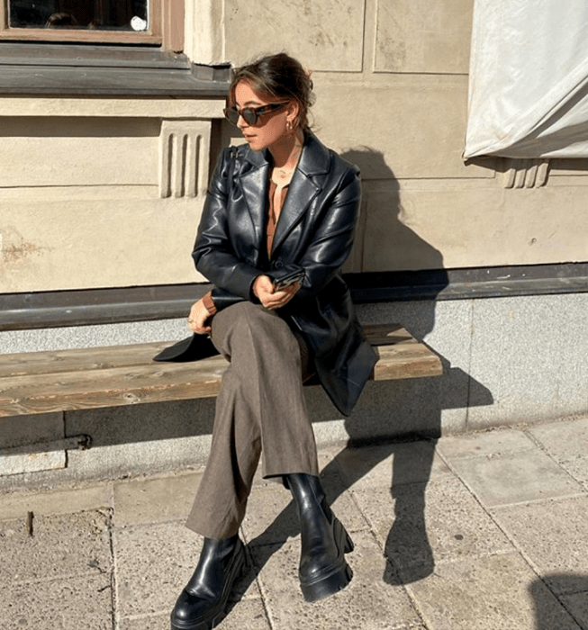 mujer de cabello castaño claro usando gafas de sol, blusa marrón, abrigo de mezclilla, pantalones de vestir marrones, botas negras de plataforma