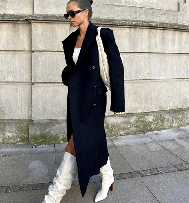 mujer de cabello corto con gafas de sol, blusa blanca, abrigo largo negro, botas blancas de tacón café y bolso blanco