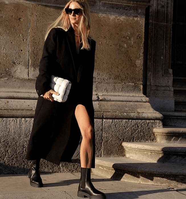 mujer rubia con gafas de sol, abrigo largo negro, botas negras de plataforma, vestido negro, bolso blanco