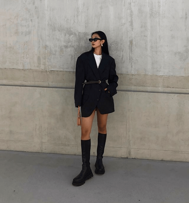 mujer de cabello oscuro con gafas de sol, camiseta blanca, abrigo oversized negro con cinturón, botas largas de plataforma gruesa