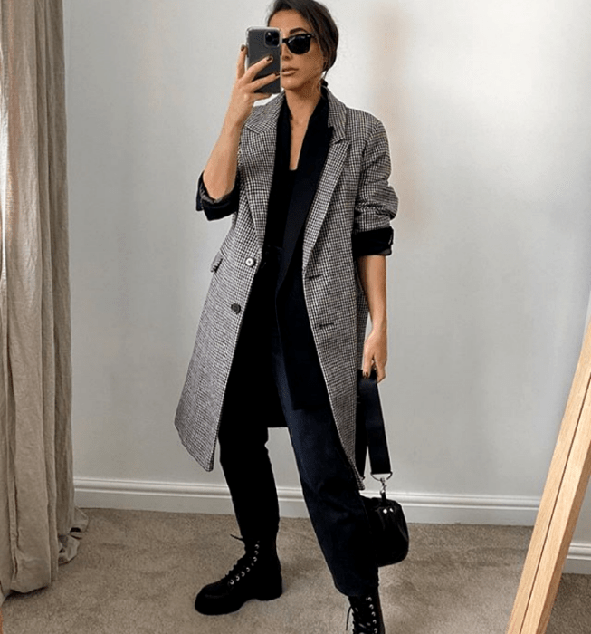 mujer de cabello castaño usando gafas de sol, blusa negra, saco negro, abrigo largo gris de cuadros, pantalones negros de vestir, botas de suela gruesa negras y mini bolso negro