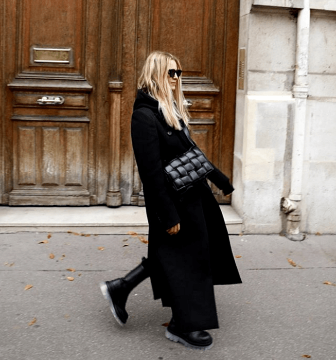 mujer rubia con gafas, vistiendo un top negro, leggings negros, abrigo largo negro, bolso negro de piel y botas de suela gruesa negras
