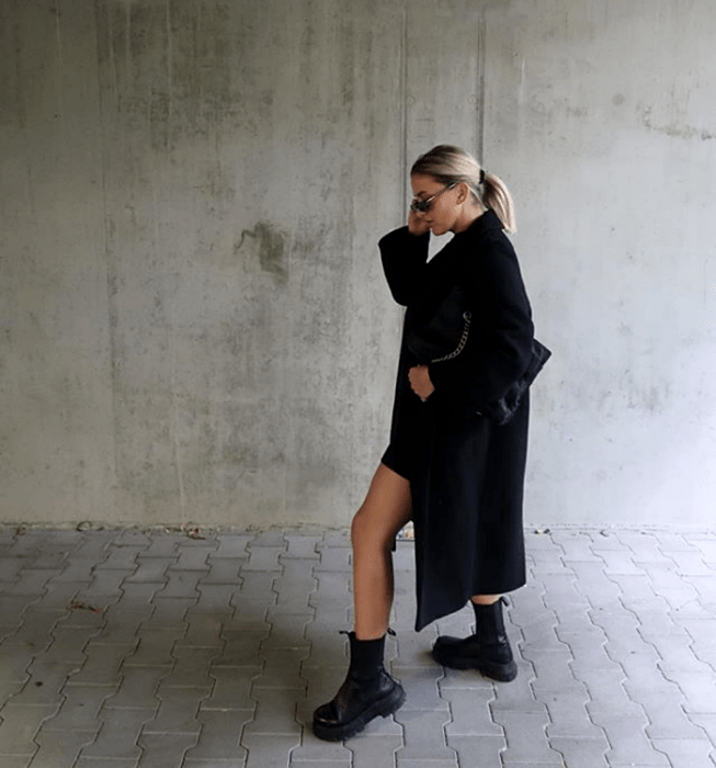 mujer rubia con gafas, usando un vestido corto negro, abrigo largo negro, botas negras de suela gruesa y bolso negro