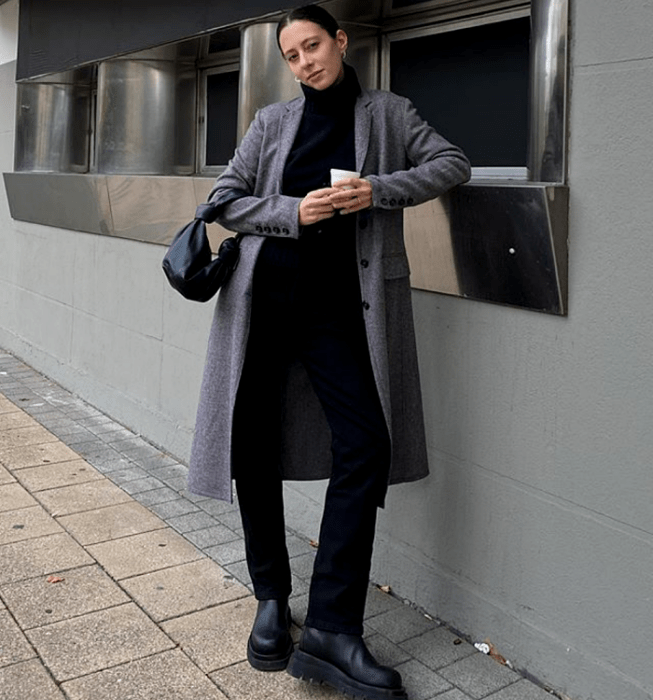 mujer de cabello castaño usando un suéter negro de cuello alto, abrigo gris largo, pantalones de vestir negros, botas de suela gruesa negras de cuero y minibolso negro
