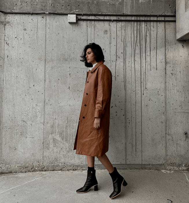 mujer de cabello castaño usando un abrigo largo de piel marrón, botas de tacón negras con suela beige