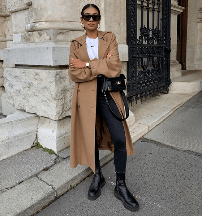mujer de cabello oscuro usando lentes de sol, camiseta blanca, abrigo largo marrón, skinny jeans negros, botas de cuero negras de suela gruesa y bolso negro
