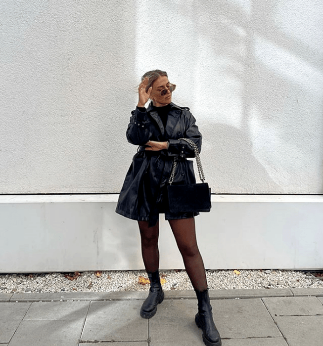 mujer rubia usando gafas, top negro de cuello alto, minifalda negra, abrigo negro de cuero, bolsa negra con cadena, medias semitransparentes y botas de suela gruesa de cuero