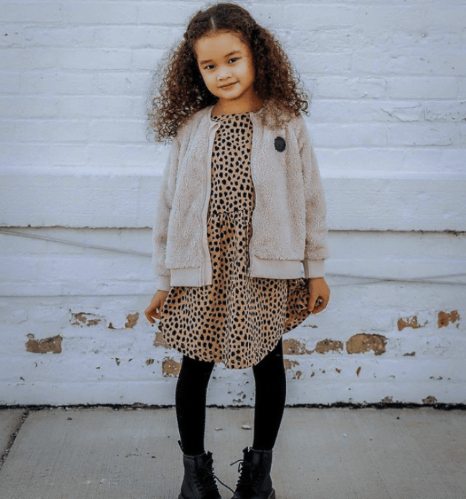 niña de cabello chino castaño usando un vestido café claro, medias negras, botines negros, suéter cárdigan beige