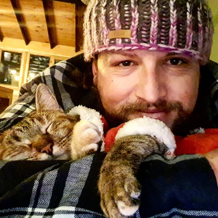 Hombre con barba con gorro tejido abraza a gato dormido con suéter rojo