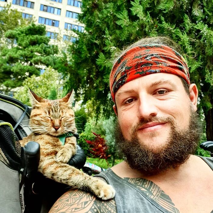 Hombre con barba con camisa de tirantes y paliacate rojo carga una mochila con gato café con rayas negras