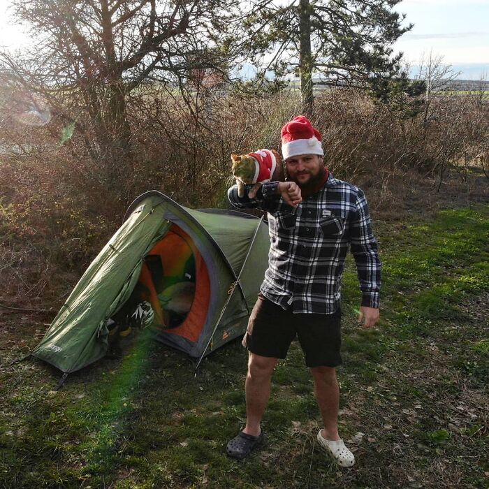 Hombre con barba vistiendo shorts y camisa a cuadros carga a un gato en su brazo con una casa de campaña verde atrás en un bosque