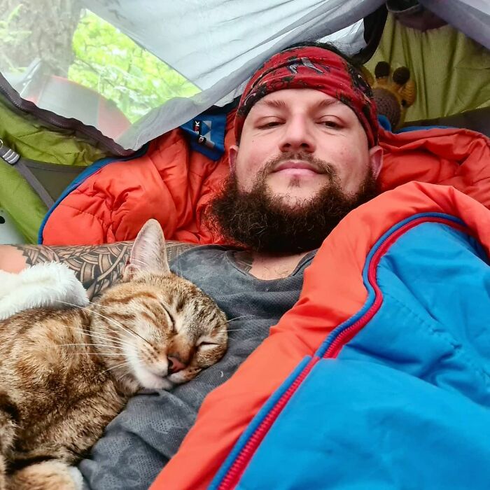 Hombre con barba y chamarra azul con interior negro tomándose selfie en casa de campaña junto a un gato café dormido en su pecho