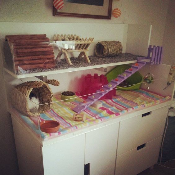 Caja para hámster colocada sobre un mueble blanco, acondicionada con camita, escaleras y muebles de madera