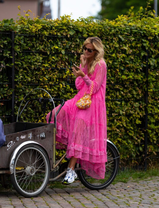 Mujer en triciclo frente a un arbusto en la calle, mirando su celular, cabello rubio, ondulado, suelto, despeinado, mediano, con vestido rosa neón de encaje y holanes hasta los tobillos, tenis skechers blancos con detalles azules, negros y verdes, bolsa de mano amarilla con cadena gruesa, lentes de sol rectangulares