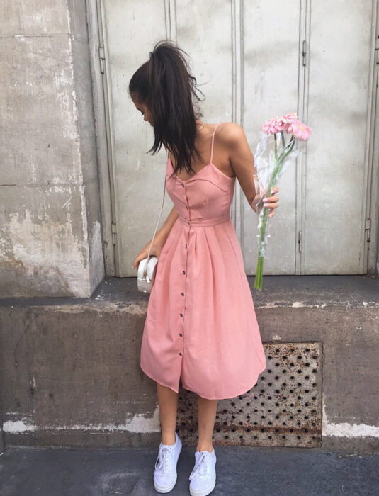 Mujer morena con vestido rosa de tirantes, botones y debajo de las rodillas, con un ramo de flores, bolsa color marfil, tenis blancos, cabello castaño, largo, lacio peinado con una cola de caballo alta