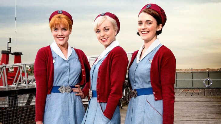 Escena de la serie Call the Midwife, trío de amigas llevando vestido azul y suéter rojo