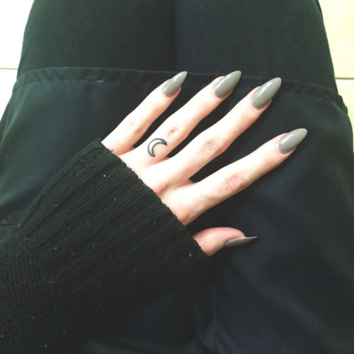 Manos cuidadas con manicura, uñas largas en forma de almendra, esmaltadas en gris verde, tatuaje de luna en el dedo corazón
