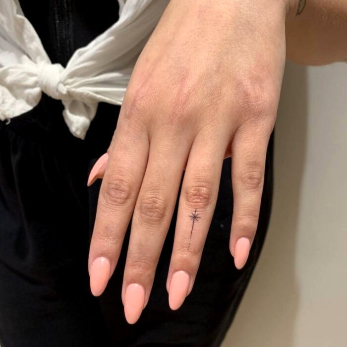 Manos bien cuidadas con manicura, uñas largas en forma de almendra, en tonos nude y rosa durazno, tatuaje de estrella fugaz en el dedo del corazón