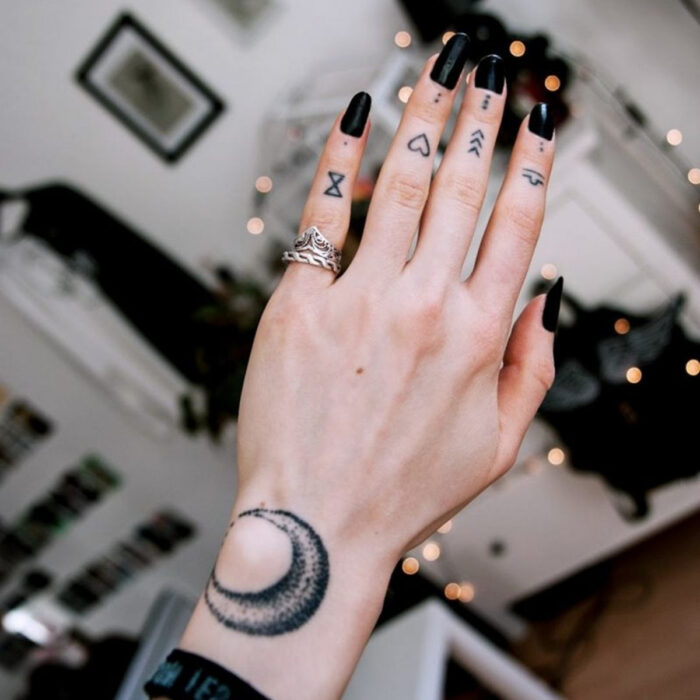 Manos cuidadas con manicura, uñas largas cuadradas en negro, dedos con tatuajes de reloj de arena, corazón y flechas, tattoo de luna en la muñeca, con anillos plateados en el meñique