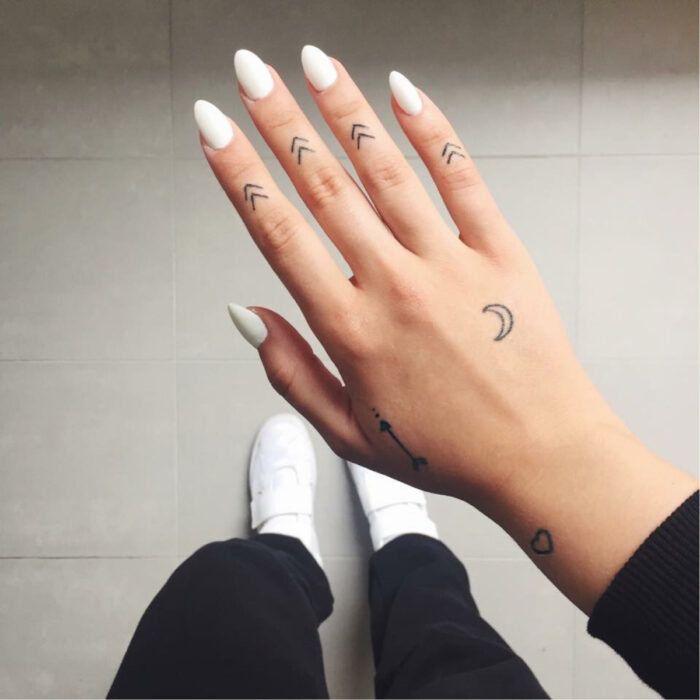 Manos elegantes con manicura, uñas largas en forma de almendra pintadas de blanco, con dedos tatuados de flechas minimalistas y otros tattoos de luna y corazón en la mano y muñeca, usando tenis blancos