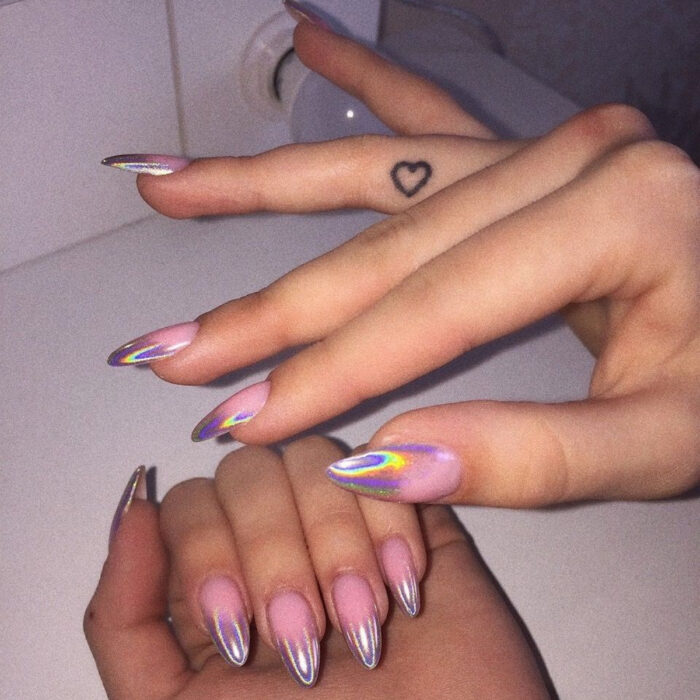 Manos elegantes con manicura, uñas largas en forma de almendra con esmalte en colores morado, verde, azul, amarillo y anaranjado, además de un tatuaje de corazón en el interior del dedo